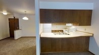 This single-floor residence offers a practical and comfortable layout designed for everyday living. The kitchen is a standout feature, showcasing rich, dark wood cabinetry that extends from counter height to the ceiling, providing generous storage space. A curved breakfast bar with a light countertop serves as the centerpiece, housing a double-basin sink and offering a natural divide between the kitchen and the adjoining living area. White appliances, including a refrigerator, electric range, and under-cabinet range hood, complement the clean, functional aesthetic of the kitchen. The space flows openly into the carpeted living area, where warm-toned, plush carpeting extends throughout, creating a cozy and inviting atmosphere. A pendant light fixture with a classic dome shade hangs in the adjacent room, adding a touch of warmth to the overall ambiance. The layout is designed to maximize the sense of openness between the kitchen and living spaces, making it well-suited for both everyday routines and casual entertaining. The consistent use of neutral tones throughout ties the space together in a cohesive and understated design.