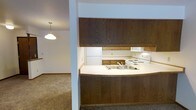 This single-floor residence offers a practical and comfortable layout designed for everyday living. The kitchen is a standout feature, showcasing rich, dark wood cabinetry that extends from counter height to the ceiling, providing generous storage space. A curved breakfast bar with a light countertop serves as the centerpiece, housing a double-basin sink and offering a natural divide between the kitchen and the adjoining living area. White appliances, including a refrigerator, electric range, and under-cabinet range hood, complement the clean, functional aesthetic of the kitchen. The space flows openly into the carpeted living area, where warm-toned, plush carpeting extends throughout, creating a cozy and inviting atmosphere. A pendant light fixture with a classic dome shade hangs in the adjacent room, adding a touch of warmth to the overall ambiance. The layout is designed to maximize the sense of openness between the kitchen and living spaces, making it well-suited for both everyday routines and casual entertaining. The consistent use of neutral tones throughout ties the space together in a cohesive and understated design.