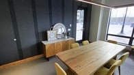 This single-floor space makes a strong visual impression through its thoughtful blend of bold design and natural materials. The interior is anchored by a dramatic accent wall finished in deep charcoal with textured vertical stripes, lending the space a modern, upscale character. A substantial live-edge wood dining table serves as a central focal point, paired with mustard yellow upholstered chairs that introduce a warm, contrasting pop of color against the darker surroundings. A beautifully crafted herringbone-patterned wood sideboard with a marble top adds refined detail, styled with decorative accents including a round metallic mirror and a classic clock. Natural light filters in through a glass-paneled door that offers a view to the exterior, brightening the space and providing a sense of connection to the outside. The flooring features a neutral carpet that grounds the room while complementing the rich wood tones throughout. Overhead, sleek linear pendant lighting runs the length of the table, enhancing both functionality and aesthetic appeal. The overall ambiance is contemporary and polished, balancing bold design choices with warm, organic textures to create an inviting and visually dynamic environment.