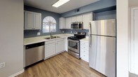 This single-story space showcases a thoughtfully designed kitchen that serves as a central highlight. The kitchen features crisp white cabinetry paired with light countertops, creating a bright and clean aesthetic against the muted blue-gray walls. Stainless steel appliances, including a refrigerator, electric range, built-in microwave, and dishwasher, lend a modern, cohesive look to the space. The double-basin sink is positioned beneath an arched window dressed with a white pleated shade, allowing natural light to filter in and illuminate the workspace. The warm wood-look flooring grounds the kitchen and adds a welcoming contrast to the cooler wall tones. The layout is designed efficiently in an L-shape, maximizing counter space and storage while maintaining an open, accessible flow. Overhead lighting ensures the space remains well-lit throughout. The overall ambiance is practical yet inviting, combining classic white finishes with contemporary stainless steel accents to create a kitchen that is both functional and visually appealing.