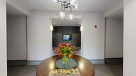 Located at 200 Perkins Dr in Chapel Hill, NC, this thoughtfully designed community space offers a refined and contemporary atmosphere across a single expansive floor. The open lobby serves as the heart of the space, anchored by bold red leather armchairs, a striped area rug, and a large-format art piece that adds visual drama. A curved accent wall with built-in display niches and a mounted flat-screen TV lends architectural interest, while large glass entry doors invite abundant natural light. A central round wood table with a modern globe chandelier above creates an inviting focal point near the reception area. The kitchen is sleek and functional, outfitted with white cabinetry, dark granite countertops, stainless steel appliances, a large island with bar seating, and pendant lighting — ideal for casual gatherings. The private dining room features a dark oval table surrounded by upholstered chairs, an open metal shelving display unit, a glass whiteboard, and a contemporary multi-globe chandelier set within a tray ceiling. Glass partition walls connect the dining room to the main lobby, maintaining visual openness throughout. Outside, a landscaped patio courtyard with wooden tables, umbrella stands, and lush greenery provides a serene setting for outdoor relaxation.