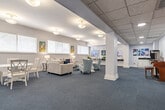 Located at Kings Reserve in Memphis, TN, this single-level community lounge provides a welcoming and thoughtfully designed gathering space. The layout is expansive, accommodating multiple functional zones that flow seamlessly into one another across the open floor plan. Blue-gray carpeting anchors the interior, complementing the soft white walls and creating a cohesive, calming aesthetic throughout.

The main lounge area features a collection of plush seating, including upholstered sofas and curved accent chairs in neutral tones accented with soft lavender and blush pillows. Rounded side tables and a glass-top coffee table add refined detail. Natural light filters in through multiple horizontal windows dressed with white blinds, and colorful artwork adorns the walls, contributing warmth and personality to the space.

A dedicated dining area with white round tables and cross-back chairs provides an intimate setting for smaller gatherings. Adjacent to this, a built-in refreshment counter with dark countertops, white cabinetry, and beverage service equipment supports community events and casual use.

A wall-mounted television, a glass-doored bookcase, and French doors leading to adjacent areas complete the space, reinforcing its role as a versatile, well-appointed community hub.