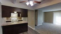 This single-floor space features an open-concept layout that connects the kitchen and living area in a functional and cohesive design. The kitchen is outfitted with dark espresso-toned cabinetry that contrasts with neutral beige walls, creating a defined visual anchor within the open floor plan. A laminate countertop peninsula serves as both a workspace and a natural divider between the kitchen and living areas. The kitchen includes standard white appliances — an electric range with coil burners and a full-size refrigerator — alongside an under-cabinet range hood. A single-basin sink is integrated into the peninsula for convenience. Hardwood-style flooring runs through the kitchen area, transitioning smoothly into plush carpeting that extends throughout the living room. The living space is generously proportioned, offering a comfortable, open feel. A ceiling fan with an integrated light fixture is centrally mounted above a subtle tray ceiling detail, adding dimension overhead. Neutral-toned walls throughout keep the space feeling bright and versatile. A doorway visible from the main living area suggests additional rooms beyond, and standard white trim and baseboards complete the clean, straightforward aesthetic of the interior.