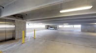 This single-level covered parking structure is built with a utilitarian yet functional design, constructed from durable concrete block walls and a solid concrete ceiling supported by prominent structural beams. The floor plan offers a generous open layout with clearly marked parking stalls, allowing for straightforward vehicle maneuvering and access. Bright fluorescent strip lighting is mounted along the ceiling, ensuring consistent visibility throughout the space. Yellow safety bollards are strategically positioned within the structure to protect both vehicles and pedestrians from potential collisions. Open horizontal gaps along the perimeter walls allow for natural light and ventilation to filter into the interior, giving the space an airy feel while maintaining shelter from the elements. Accessibility is considered, as indicated by designated accessible parking signage visible within the facility. The smooth concrete floor is divided into distinct stalls by painted line markings, supporting orderly and efficient use of the available parking area. The overall ambiance is clean, well-maintained, and practical, making this a reliable and straightforward parking facility suited to serve the needs of a commercial or residential development.