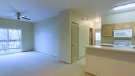 This single-floor residence offers a clean, functional layout designed for comfortable everyday living. The open-concept design connects the living area to the kitchen, creating a sense of spaciousness and ease of movement throughout the home. Soft, neutral wall tones in warm beige and light gray run throughout the space, contributing to a calm and welcoming ambiance. Wall-to-wall carpeting in the living area provides a cozy underfoot feel and helps define the space. The kitchen features a raised breakfast bar counter that overlooks the main living area, along with warm wood-toned cabinetry and efficient task lighting overhead. A pass-through view from the kitchen into adjacent areas enhances the open, airy feel of the home. Interior doors with classic six-panel detailing and wood-trimmed frames add a traditional architectural touch. Ceiling-mounted light fixtures provide consistent illumination throughout, while visible air vents indicate a central HVAC system for year-round climate control. The layout includes additional rooms accessible through hallways, hinting at a thoughtfully organized floor plan that balances shared and private living spaces. Overall, the residence presents a tidy, move-in-ready environment with a straightforward and practical design.