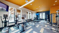 This fitness center in Scott, Louisiana, delivers a dynamic and energizing workout environment designed to motivate and perform. The space is defined by a bold, contemporary aesthetic — a striking orange ceiling contrasts with deep navy blue accent walls, while a large "Train Hard" motivational mural adds an unmistakable sense of purpose to the room. Full-length mirrors along one wall visually expand the space and allow users to monitor their form during exercises.

The flooring consists of patterned modular carpet tiles in shades of blue, gray, and cream, providing both visual interest and practical cushioning underfoot. Large windows line two walls, flooding the interior with abundant natural light and offering views of the surrounding outdoor areas.

The cardio zone is well-appointed with multiple treadmills, elliptical trainers, a stair climber, and a stationary bike, each equipped with individual digital consoles. A wall-mounted flat-screen television provides entertainment during workouts. The strength training area features a cable machine, a power rack, barbells, weight plates, and a full dumbbell rack covering a wide range of weights. Medicine balls and additional free weights round out the equipment selection.

Overhead linear pendant lighting supplements the natural light, ensuring the space remains bright and well-illuminated at all hours. The overall design balances high-energy aesthetics with functional layout, making this fitness center a comprehensive and inviting facility.