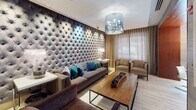This elegantly designed single-floor space makes an immediate impression with its standout floor-to-ceiling tufted velvet accent wall, which anchors the main lounge area in understated luxury. The lounge is furnished with a plush gray sofa accented by teal and blue throw pillows, complemented by a live-edge wood coffee table and coordinating armchairs. A striking cylindrical chandelier with crystal detailing overhead adds a glamorous focal point to the room. Warm, sheer drapery softens the perimeter walls and allows diffused natural light to filter through, creating an inviting and comfortable atmosphere. Light-toned wood-effect tile flooring flows seamlessly throughout, unifying the various zones of the space. Rich wood-paneled structural columns provide architectural definition, separating the lounge from an adjacent seating area bathed in warm accent lighting, where a textured golden wall adds depth and character. A well-stocked built-in bookshelf further enhances the space's cultivated aesthetic. Beyond the lounge, a bright dining area with a round table is visible through an open passage, offering views of a lively streetscape outside. The overall ambiance is polished and contemporary, blending luxurious textures with thoughtful design details throughout.