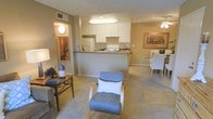 This single-floor residence showcases an open-concept design that seamlessly connects the living, kitchen, and dining areas into one cohesive, flowing space. The living area features neutral-toned walls and plush carpeting underfoot, creating a warm and inviting atmosphere. A cozy seating arrangement, including a modern accent chair with a cushioned back, anchors the room alongside a decorative console table.

The kitchen is positioned centrally, defined by a raised breakfast bar that separates it from the living space without disrupting the open feel. White cabinetry with ample storage lines the walls, complemented by a white appliance suite including a refrigerator and range. Dark countertops provide a subtle contrast to the bright cabinetry above.

Adjacent to the kitchen, the dining area comfortably accommodates a full table and chair set, lit softly by a ceiling fan with integrated lighting. Framed artwork adorns the walls throughout, adding character to the neutral palette. A hallway off the living room leads to additional private spaces, with wall-mounted lighting guiding the way. Overall, the layout prioritizes functionality while maintaining a comfortable, cohesive aesthetic.