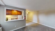 This single-floor unit is thoughtfully laid out to maximize functionality across its living spaces. The open living area is finished with neutral-toned carpet and painted walls in soft beige and gray, creating a clean, understated aesthetic throughout. A ceiling fan with a light fixture provides overhead illumination and air circulation to the main living space.

The kitchen is accessed via a pass-through window framed in white trim, which visually connects it to the living area. Warm honey-toned wood cabinetry lines the upper walls of the kitchen, complemented by a dark countertop and a stainless sink. A compact white refrigerator is also visible within the kitchen area.

The bedroom offers a modestly sized, carpeted space with neutral walls and ample room for standard furnishings. A second carpeted room provides additional flexible space suitable for various uses.

The bathroom features white subway tile accents along the walls, wood-look flooring, a white vanity with an integrated sink, a wall-mounted mirror, and a shower-tub combination enclosed by a decorative curtain. White trim and clean finishes maintain a cohesive, practical look throughout the unit.