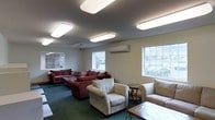 This single-floor space serves as a comfortable and well-appointed communal lounge designed for relaxation and social interaction. The room is furnished with an ample arrangement of seating, including a large red sectional sofa, cream-colored armchairs, and a matching sofa, offering a variety of comfortable options for occupants and guests alike. A combination of wooden side tables and a coffee table adds functional surface space throughout the seating area.

The room is illuminated by multiple large, flush-mounted rectangular panel lights fixed to the ceiling, ensuring bright and even lighting throughout the space. A wall-mounted split-system air conditioning unit provides efficient climate control, enhancing year-round comfort. The green carpeted flooring adds warmth underfoot and contributes to the cozy, inviting atmosphere of the room.

Natural light filters in through several windows positioned along the walls, offering views of the surrounding outdoor greenery and bringing a sense of openness to the interior. The neutral, cream-toned walls complement the overall warm and welcoming ambiance. This thoughtfully arranged lounge space is well-suited for use as a community gathering area, waiting room, or shared recreational space.