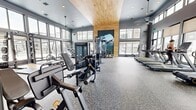 Located at 3001 S 21st Ave in Bozeman, Montana, this contemporary fitness center offers a generously sized, single-floor layout thoughtfully divided into distinct workout zones. The main exercise floor is defined by soaring double-height ceilings clad in warm wood paneling, floor-to-ceiling windows along multiple walls, and speckled rubber flooring built for heavy use. Rows of treadmills, ellipticals, stair climbers, and rowing machines line the perimeter, each bathed in abundant natural light and framed by views of the surrounding Bozeman community. A dedicated strength area houses a comprehensive cable and functional training machine, a fully stocked dumbbell and kettlebell rack, stability balls, and adjustable benches, all reflected in large wall-mounted mirrors beneath mounted flat-screen displays. A separate studio room provides a quieter environment for cycling classes and focused training, featuring stationary bikes, soft carpet flooring, roller shades for light control, and a vibrant Montana landscape mural. Throughout the facility, ceiling fans maintain airflow, recessed lighting supplements natural illumination, and nature-themed murals evoke the rugged outdoor character of the region, creating an energizing and visually cohesive workout environment.