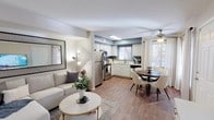 This single-floor residence offers a seamlessly connected open-concept layout, blending the living, dining, and kitchen areas into one cohesive and functional living space. Warm hardwood-style flooring runs throughout, providing a consistent and inviting foundation across all areas.

The kitchen is well-appointed with stainless steel appliances, including a refrigerator and range, complemented by crisp white cabinetry and light countertops. A skylight overhead floods the kitchen with natural light, enhancing the airy atmosphere. The adjoining dining area comfortably accommodates a round table and seating, positioned near large windows dressed with white curtains that allow generous natural light to filter in.

The living area is tastefully styled with a neutral-toned sofa, a round coffee table with gold accents, and a large decorative mirror that amplifies the sense of space. A ceiling fan is installed overhead, adding both comfort and a functional design element. Soft, accent wall sconces contribute to a warm and welcoming ambiance.

The overall aesthetic is modern and clean, characterized by a neutral color palette of whites, soft grays, and muted blue accent walls. The open flow between spaces makes the residence feel expansive and well-suited for both everyday living and entertaining.