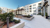 This thoughtfully designed amenity space serves as a vibrant communal hub within a contemporary multi-story residential community. At its centerpiece is a resort-style swimming pool with in-water lounge chairs and a shallow sun shelf, all framed by a clean concrete deck and the surrounding building's modern stucco and glass facade. Adjacent to the pool, a spacious open-air courtyard features manicured artificial turf, herringbone-patterned paver walkways, and an assortment of seating options — from hanging egg chairs to modular outdoor furniture — providing multiple areas for relaxation. Tall palm trees and curated landscaping add a warm, resort-like atmosphere throughout the outdoor areas. A covered outdoor kitchen equipped with a stainless steel grill station and sink sits beneath a pergola-style canopy, offering a dedicated space for outdoor cooking and entertaining. The covered indoor-outdoor lounge area is a standout feature, with large-format tile flooring, string lighting, ceiling fans, and distinctive rope-suspended swings and hanging basket chairs. A wet bar with cabinetry and sink, along with a wall-mounted television and long communal dining table, make this area well-suited for gatherings. The seamless flow between indoor and outdoor zones, combined with abundant natural light, creates an inviting and functional amenity space.