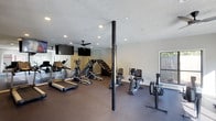 This fitness center offers a spacious, open layout designed to accommodate a comprehensive cardio and strength training experience. The room is finished with dark, low-pile carpet underfoot, providing a practical and durable surface suited for exercise equipment. Recessed lighting throughout the textured ceiling ensures the space is well-lit, while ceiling fans promote air circulation during workouts.

The facility is outfitted with a variety of cardio machines, including treadmills, elliptical trainers, a stair climber, and stationary bikes, arranged to allow comfortable movement between stations. Wall-mounted flat-screen televisions are positioned for easy viewing from multiple machines, enhancing the workout experience. A large mirror along one wall visually expands the space and allows users to monitor their form.

Structural support columns are integrated into the floor plan, adding an industrial character to the room. Natural light filters in through a window on one side, offering a view of a landscaped outdoor area. The neutral wall color and clean design create a focused, distraction-free environment. Overall, this fitness center is thoughtfully designed to provide a fully functional and comfortable workout facility.