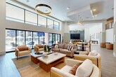 Located at 14965 W Encanto Blvd in Goodyear, Arizona, this generously sized single-floor community clubhouse offers an impressive and well-appointed social space designed for both relaxation and recreation. The space is defined by its soaring double-height ceilings, crisp white walls, and expansive floor-to-ceiling windows that flood the interior with abundant natural light while offering direct views of the outdoor pool and patio area beyond.

The living area is anchored by comfortable lounge seating arranged around neutral-toned coffee tables atop patterned area rugs, all set on warm wood-look tile flooring. A bold circular gold pendant light serves as a focal point overhead, complementing the modern yet inviting aesthetic. The entertaining zone features a full-size pool table and a shuffleboard table, both set beneath elegant chandelier lighting, alongside a large flat-screen TV mounted against a textured stone accent wall.

A dedicated game nook showcases a wall-mounted board game display alongside casual seating, while a dining and collaborative area provides long communal tables with a mix of leather and upholstered chairs. Throughout, decorative wall art, woven textures, and curated furnishings contribute to a cohesive, resort-inspired atmosphere. The seamless connection between the interior and the outdoor pool deck makes this a dynamic and vibrant communal hub.