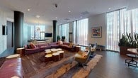This thoughtfully designed lounge offers a dynamic, multi-zone layout that balances entertainment, relaxation, and social interaction. The space is anchored by a sprawling seating area furnished with deep burgundy leather sectional sofas, colorful accent pillows, and a collection of unique hexagonal wood coffee tables atop a richly textured area rug. Swivel lounge chairs and framed artwork contribute to the sophisticated, eclectic atmosphere.

A dedicated game area hosts a full-sized billiards table beneath a striking starburst chandelier, while floor-to-ceiling sheer-draped windows flood the zone with natural light. A sleek flat-screen TV is mounted against a textured stone accent wall, creating a media-focused nook with additional seating.

The kitchen and bar area is a visual highlight, featuring bold teal cabinetry with high-gloss finishes, a long dark countertop bar, and a decorative rope installation overhead, adding an artistic, nautical touch. Warm hardwood flooring flows throughout, unifying each zone cohesively. Dark cylindrical columns and recessed lighting complement the contemporary industrial aesthetic, while abundant natural light and curated greenery enhance the overall inviting ambiance.