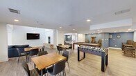 Located at 8503 N Tryon St in Charlotte, NC, this single-floor clubhouse offers a generously sized, open-concept layout designed for comfort and community engagement. The entrance foyer welcomes visitors with soaring vaulted ceilings, a dramatic multi-pendant chandelier, a skylight that floods the space with natural light, and a striking accent wall in deep teal blue. Large floor-to-ceiling windows frame views of the lush landscaped grounds outside, further enhancing the bright and airy atmosphere throughout.

The expansive living room serves as the heart of the space, anchored by a floor-to-ceiling stacked stone fireplace and complemented by double-height ceilings with pendant lighting. Light wood-tone flooring runs throughout, unifying the open layout. A wall-mounted flat-screen television and comfortable seating arrangements make this area both functional and inviting.

The dining area seamlessly connects to the living space and extends into a game room equipped with a billiard table, foosball table, and casual seating—ideal for social gatherings. A separate, more intimate room provides a quiet setting with its own wall-mounted display screen and natural light, suitable for meetings or focused activities. The overall design balances modern aesthetics with a warm, community-focused ambiance.