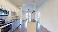 This single-floor residence showcases a thoughtfully designed open-concept layout where the kitchen and living area flow seamlessly together. The kitchen is a standout feature, combining white upper shaker-style cabinetry with slate-blue lower cabinets and dark granite countertops. A large central island with matching dark granite provides additional prep and dining space, illuminated overhead by two sleek black-and-gold pendant lights. Stainless steel appliances, including a French door refrigerator, electric range, built-in microwave, and dishwasher, complement the subway tile backsplash. Rich hardwood-style flooring extends throughout the main living area, anchored by recessed lighting and a ceiling fan. Expansive windows and a set of glass panel doors allow generous natural light to fill the living space while offering outdoor views. The bedroom is finished with plush neutral carpet and a trio of large windows that continue the theme of bright, airy interiors. The bathroom features wood-style flooring, a soaking tub surrounded by large-format marble-look tile, and a vanity with dark granite countertops, creating a clean, polished aesthetic throughout.