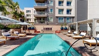 Located at 98 N 1st St in the heart of downtown San Jose, California, this thoughtfully designed outdoor amenity space offers a resort-like retreat within an urban residential setting. The centerpiece of the space is a generously sized swimming pool with crystal-clear water, flanked by a sun deck lined with lounge chairs and shade umbrellas — ideal for relaxing on sunny California days. Adjacent to the pool, a spa provides an additional space for leisure and unwinding.

A dedicated fire pit lounge area features a sleek, glass-enclosed gas fire table set within a wraparound built-in seating arrangement, accented with plush cushions and decorative pillows, creating a welcoming gathering space for evening relaxation. Raised planter beds throughout the space are filled with a diverse mix of tropical and Mediterranean-inspired greenery, including towering palm trees, agaves, ornamental grasses, and flowering shrubs, contributing to a vibrant and lush atmosphere.

The hardscape design incorporates stone-paved walkways and tiered landscaping with accessible ramps and railings, blending functionality with aesthetic appeal. The multi-story residential buildings surrounding the courtyard provide a sense of enclosure and community, while still allowing ample natural light to flood the space throughout the day.