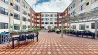 Located at 900 Luxor Ln in Norristown, PA, this outdoor courtyard space is a well-appointed amenity area nestled within a modern multi-story building complex. The architecture features a striking combination of bold red brick and cream-toned panels, with large windows lining each floor and providing an attractive backdrop to the courtyard below.

The courtyard is anchored by a patterned brick paver plaza in warm red and tan tones, creating a visually dynamic surface throughout the space. A pergola with string lighting serves as a shaded gathering point, complemented by multiple outdoor dining tables and chairs ideal for social gatherings. A gas grill station makes the area well-suited for outdoor cooking and entertaining.

A standout feature of the courtyard is the full-length bocce ball court, surfaced in turf and framed by clean metal borders, offering recreational activity for residents or visitors. Adjacent to the court, a generous turf lawn provides additional open green space for relaxation or casual use.

Raised garden planters filled with ornamental grasses, shrubs, and slender trees line the perimeter, introducing natural greenery and softening the architectural edges of the surrounding building. A lounge seating area with all-weather furniture adds a comfortable retreat within the courtyard. The space is open to the sky, allowing ample natural light throughout the day.