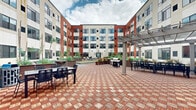Located at 900 Luxor Ln in Norristown, PA, this outdoor courtyard space is a well-appointed amenity area nestled within a modern multi-story building complex. The architecture features a striking combination of bold red brick and cream-toned panels, with large windows lining each floor and providing an attractive backdrop to the courtyard below.

The courtyard is anchored by a patterned brick paver plaza in warm red and tan tones, creating a visually dynamic surface throughout the space. A pergola with string lighting serves as a shaded gathering point, complemented by multiple outdoor dining tables and chairs ideal for social gatherings. A gas grill station makes the area well-suited for outdoor cooking and entertaining.

A standout feature of the courtyard is the full-length bocce ball court, surfaced in turf and framed by clean metal borders, offering recreational activity for residents or visitors. Adjacent to the court, a generous turf lawn provides additional open green space for relaxation or casual use.

Raised garden planters filled with ornamental grasses, shrubs, and slender trees line the perimeter, introducing natural greenery and softening the architectural edges of the surrounding building. A lounge seating area with all-weather furniture adds a comfortable retreat within the courtyard. The space is open to the sky, allowing ample natural light throughout the day.