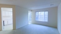 This single-floor space presents a clean and open layout defined by neutral tones and an abundance of natural light. The main living area is finished with soft, wall-to-wall carpeting in a light neutral shade, contributing to a comfortable and quiet atmosphere underfoot. Smooth white walls are paired with a textured ceiling, giving the room a simple yet polished appearance.

A standout feature of the space is the large multi-pane window along one wall, which draws in generous amounts of natural light and frames views of surrounding trees, creating a pleasant connection to the outdoors. Adjacent to the window, a sliding glass door provides direct access to an outdoor balcony or patio area, further enhancing the sense of openness and allowing for additional natural light to enter the room.

A doorway leads to an adjoining room or hallway, suggesting a functional and connected floor plan. The overall ambiance of the space is bright, airy, and neutral, making it a versatile canvas well-suited for a variety of furnishing styles and layouts. The wooded outdoor views visible through the windows add a calm, natural quality to the interior environment.