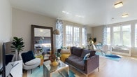 This thoughtfully designed single-floor residence offers an open-concept layout that seamlessly connects the living, dining, and kitchen areas into one cohesive, functional space. The living area is anchored by a plush charcoal sofa accented with colorful throw pillows and a striking geometric glass coffee table, all resting atop a soft teal area rug that adds a vibrant pop of color to the light hardwood flooring. A large floor-length mirror with a dark wood frame amplifies the sense of space, while a sleek arc floor lamp provides warm ambient lighting. Botanical-patterned drapery panels frame the tall, multi-pane windows that flood the interior with generous natural light and offer views to the outdoors. The dining area features a round glass table paired with modern white chairs, creating an airy and elegant setting for meals. Adjacent to the living space, the kitchen is finished with dark cabinetry, stainless steel appliances including an over-the-range microwave, and ample counter space, offering a modern and efficient workspace. Smooth white ceilings and neutral wall tones throughout lend the space a clean, contemporary aesthetic that is both inviting and versatile.