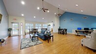 Located at 20 Frontier Ridge Ct in Staunton, Virginia, this single-floor community space offers an open, welcoming layout designed for shared gatherings and everyday comfort. The interior is immediately distinguished by dramatic vaulted ceilings that rise sharply overhead, lending an airy, expansive feel throughout. Dual ceiling fans and recessed lighting ensure the space remains bright and comfortable at all times.

Warm hardwood-style flooring runs continuously across the entire floor, unifying the various functional zones. The central living area features a comfortable seating arrangement with a blue sofa, a patterned area rug, and a wall-mounted flat-screen television set against a bold slate-blue accent wall — a standout architectural detail that anchors the space visually.

Multiple dining table sets are positioned across the room, accommodating a variety of group sizes. Large windows along one wall flood the dining areas with natural light and offer views of the surrounding neighborhood. A dedicated workstation with a computer desk provides a functional nook for individual use.

A well-appointed kitchen features warm wood cabinetry, ample counter space, and full appliances including a range, refrigerator, microwave, and dishwasher. Near the entrance, a welcoming reception area includes comfortable armchairs and service windows. Glass-paned entry doors complete the bright, community-oriented atmosphere of this versatile space.