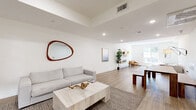 Located at 418 N Norton Ave in Los Angeles, this single-floor space offers a thoughtfully designed open-plan layout that maximizes its footprint with a seamless, airy feel. The interior is anchored by light wood-toned flooring that runs continuously throughout, complementing the crisp white walls and creating a warm, contemporary atmosphere.

The living area is generously proportioned, featuring recessed ceiling lighting alongside stylish wall-mounted swing-arm lamps that provide both functional and ambient illumination. A tray ceiling detail adds subtle architectural interest overhead. Natural light pours in through multiple windows, including a large gridded window near the entry that frames views of the surrounding greenery.

The space flows effortlessly from a dedicated lounge zone — complete with a deep sectional sofa, organic-shaped coffee table, and woven area rug — into a secondary gathering area with a rustic wood dining table and bench seating. Curated wall art, an asymmetric wood-framed mirror, and open shelving with decorative accents contribute to the refined, modern-minimalist character of the room.

The entry area is clean and welcoming, with a solid door, a framed window, and a potted olive tree that adds a natural, organic touch. Overall, the space conveys a calm, sophisticated ambiance well-suited to both relaxed living and social gatherings.