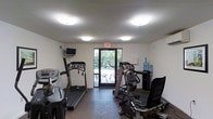 This single-floor fitness room offers a practical and well-organized space for cardiovascular exercise. The room features smooth white walls and a flat ceiling fitted with multiple flush-mount light fixtures that provide consistent, even illumination throughout. A glass-paneled door along the back wall opens directly to an outdoor patio area, allowing natural light to filter in and providing views of surrounding greenery, which adds an open, airy quality to the space.

The room is equipped with several cardio machines, including two treadmills positioned along one side, a stationary upright bike, an elliptical trainer, and what appears to be a recumbent-style exercise chair. A small wall-mounted television is installed near the entrance, offering entertainment during workouts. A water cooler with a large refill jug sits in the corner alongside a multi-tiered storage rack, ensuring hydration and organizational needs are met. The flooring transitions from a darker material near the entrance to a lighter surface toward the patio door. An illuminated emergency exit sign is centrally positioned above the back door, meeting safety requirements. Overall, the space is functional, clean, and practical for everyday fitness use.