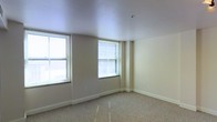 This single-floor space offers a clean and versatile layout with a neutral, understated aesthetic throughout. The main room is open and generously proportioned, featuring smooth white ceilings that enhance the sense of openness and reflect natural light effectively. Multiple double-hung windows fitted with horizontal blinds line the walls, allowing ample daylight to fill the interior and creating a bright, welcoming atmosphere. The walls are finished in a soft, warm off-white tone, complementing the light-colored speckled floor that adds subtle texture underfoot. Structural column elements are seamlessly integrated into the corners of the room, contributing to the building's architectural character without disrupting the flow of the space. Clean white baseboards provide a crisp finish along the perimeter. The overall layout is unencumbered and adaptable, making it well-suited for a range of configurations and uses. The space presents a blank canvas with solid bones — neutral finishes, good natural light, and a straightforward, functional design that can be easily personalized or outfitted to meet specific needs.