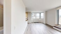 Located at 401 East 34th Street, Unit S18J in Manhattan, this 570-square-foot, one-bedroom, one-bathroom apartment delivers a practical and well-appointed urban living experience. The open-plan kitchen and living area serves as the heart of the unit, featuring rich dark wood cabinetry, dark granite countertops, a glass tile backsplash, and stainless steel appliances including a refrigerator, microwave, and range. Light hardwood-style flooring runs throughout the apartment, creating a cohesive and clean aesthetic. The living area is bathed in natural light through large floor-to-ceiling sliding glass doors that open directly onto a shared outdoor terrace, offering open city views. The 160-square-foot primary bedroom features a window with neighborhood views and direct access to a dedicated closet. A separate hallway closet provides additional storage. The entry hallway includes a building intercom system. The overall design is characterized by neutral tones, clean white walls, and an efficient layout that maximizes every square foot of the 570-square-foot floor plan.