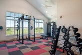This fitness center offers a spacious and energizing environment designed to support a comprehensive workout experience. The space is defined by its dramatic vaulted ceiling, which rises steeply to a peak, creating an open and airy atmosphere enhanced by recessed lighting and dual ceiling fans that ensure comfort during workouts. Large windows along the walls flood the interior with natural light and provide views of the surrounding greenery, contributing to an uplifting ambiance.

The flooring consists of bold, multi-colored modular carpet tiles in shades of red, orange, tan, and gray, arranged in a striking checkerboard-inspired pattern that adds a dynamic visual energy to the space. A large wall-mounted mirror along one side expands the sense of space and assists with form during exercise.

The cardio zone is well-appointed with a lineup of treadmills, elliptical machines, and stair climbers, each equipped with digital displays. Multiple flat-screen televisions are mounted on the walls to keep users engaged during cardio sessions. The strength training area features a multi-function power rack for functional and free weight training, alongside multiple dumbbell racks stocked with a full range of weights. A red accent wall near the entrance adds a bold architectural focal point, complementing the overall energetic design of the facility.