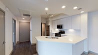 This single-floor space showcases a contemporary open-concept kitchen that serves as the central feature of the layout. The kitchen is designed with clean lines and a minimalist aesthetic, highlighted by an expansive white peninsula that includes a double stainless steel sink and ample counter space, making it ideal for both meal preparation and casual dining.

Crisp white cabinetry extends to the ceiling, providing generous storage while reinforcing the modern design theme. Stainless steel appliances, including a gas range, built-in microwave, and refrigerator, are seamlessly integrated into the layout. A track lighting fixture above the kitchen workspace offers targeted illumination, complementing the recessed ceiling lights that are evenly distributed throughout the space.

Dark hardwood-style flooring grounds the interior and creates a striking contrast against the white cabinetry and walls. A hallway visible from the kitchen leads to additional rooms, with a neutral-toned entry door and a secondary door providing clear access points. The overall ambiance is bright, clean, and functional, reflecting a well-organized modern residential design.