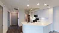 This single-floor space showcases a contemporary open-concept kitchen that serves as the central feature of the layout. The kitchen is designed with clean lines and a minimalist aesthetic, highlighted by an expansive white peninsula that includes a double stainless steel sink and ample counter space, making it ideal for both meal preparation and casual dining.

Crisp white cabinetry extends to the ceiling, providing generous storage while reinforcing the modern design theme. Stainless steel appliances, including a gas range, built-in microwave, and refrigerator, are seamlessly integrated into the layout. A track lighting fixture above the kitchen workspace offers targeted illumination, complementing the recessed ceiling lights that are evenly distributed throughout the space.

Dark hardwood-style flooring grounds the interior and creates a striking contrast against the white cabinetry and walls. A hallway visible from the kitchen leads to additional rooms, with a neutral-toned entry door and a secondary door providing clear access points. The overall ambiance is bright, clean, and functional, reflecting a well-organized modern residential design.