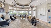 Located at 19411 Via del Mar in Tampa, FL, this single-story community clubhouse offers a well-appointed collection of shared amenity spaces designed for both relaxation and productivity. The entrance foyer greets visitors with diagonal tile flooring, arched architectural details, columned accents, and a set of glass double doors that draw in natural light and frame views of the surrounding landscape. Workstation desks flank the entry, providing convenient access points for residents. The main lounge is a standout feature, showcasing a dramatic octagonal tray ceiling with recessed lighting and crown molding. A sweeping curved bay window wall wraps around the room, offering panoramic views of the outdoor pool and lush tree line beyond. The space is furnished with plush seating, a glass coffee table, and intimate conversation areas. An adjacent sitting nook with built-in shelving and a glass-panel door provides additional pool access. A dedicated business center offers computer workstations, comfortable seating, and natural light from a side window. The fitness center is spacious and well-equipped, featuring cardio machines including treadmills and ellipticals, strength training equipment, free weights, and exercise balls. Floor-to-ceiling mirrors, rubber flooring, and ceiling fans create a functional and motivating workout environment. Together, these spaces form a cohesive, elegantly designed community hub.