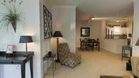 This single-story space showcases an open-concept layout that allows the living and dining areas to flow naturally into one another. The living area features plush, neutral-toned carpeting that extends throughout, contributing to a warm and cohesive aesthetic. Crown molding adds a refined architectural detail along the ceiling line, enhancing the overall sense of polish. A partial wall with a smooth, glossy white finish acts as a subtle divider between the two spaces without compromising the open feel.

The living area is accented with decorative wall art featuring an ornate scrollwork design, complemented by a classic floor lamp with a black shade. A patterned accent chair and a small side table complete the seating area with understated elegance.

The adjacent dining area accommodates a dark wood dining set with matching chairs, anchored by a framed mirror on the wall that reflects light and adds visual depth. Track lighting on the ceiling provides functional illumination throughout. Built-in shelving is visible in the background, offering additional display and storage options. Horizontal blinds on the windows allow for flexible light control. The overall ambiance is neutral, tidy, and well-appointed.