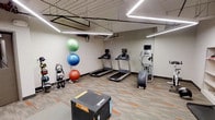 This fitness room offers a practical and well-organized space for a comprehensive workout experience. The room features an industrial-style ceiling with exposed ductwork and piping, complemented by modern linear LED strip lighting that ensures bright, even illumination throughout. The flooring consists of durable, multi-toned carpet tiles in neutral and orange accents, providing both comfort and resilience underfoot.

The cardio section is anchored by multiple treadmills equipped with individual screens, alongside an elliptical or stair-climber machine, all positioned along the walls for efficient use of space. A functional training area includes a heavy plyo box positioned centrally, ideal for jump training and conditioning exercises.

Along one wall, a rack of kettlebells in varying weights provides options for strength and functional fitness training. A colorful collection of stability and exercise balls — in green, blue, and red — adds versatility for core and balance workouts. The neutral walls and organized layout create a clean, focused environment suitable for a wide range of exercise routines. Overall, the space is efficiently designed to support both cardio and strength-based training.