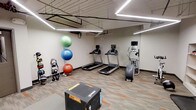 This fitness room offers a practical and well-organized space for a comprehensive workout experience. The room features an industrial-style ceiling with exposed ductwork and piping, complemented by modern linear LED strip lighting that ensures bright, even illumination throughout. The flooring consists of durable, multi-toned carpet tiles in neutral and orange accents, providing both comfort and resilience underfoot.

The cardio section is anchored by multiple treadmills equipped with individual screens, alongside an elliptical or stair-climber machine, all positioned along the walls for efficient use of space. A functional training area includes a heavy plyo box positioned centrally, ideal for jump training and conditioning exercises.

Along one wall, a rack of kettlebells in varying weights provides options for strength and functional fitness training. A colorful collection of stability and exercise balls — in green, blue, and red — adds versatility for core and balance workouts. The neutral walls and organized layout create a clean, focused environment suitable for a wide range of exercise routines. Overall, the space is efficiently designed to support both cardio and strength-based training.