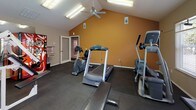 This fitness center is housed in a single-story space distinguished by vaulted ceilings that lend an open, airy feel to the room. The ceiling is outfitted with flush fluorescent light fixtures and a ceiling fan, ensuring consistent brightness and airflow throughout workouts. Neutral beige walls on one side contrast with a warm accent wall in a deep amber tone, creating a visually dynamic environment.

The rubber-tiled flooring covers the entire workout area, providing cushioning and traction suitable for a variety of exercises. Large windows with blinds line the walls, allowing natural light to fill the space and offering views of an outdoor pool area and surrounding greenery. A wall-mounted flat-screen television is positioned for easy viewing during cardio sessions.

Equipment is thoughtfully arranged throughout the room. Cardio options include a treadmill, elliptical trainers, a stair climber, and a stationary bike. A multi-station cable machine supports a full range of strength training exercises, complemented by a dedicated free weight area featuring a dumbbell rack with varied weights and an adjustable workout bench. A stability ball and a vending machine are also present. Mirrored walls on one side expand the perceived scale of the space.