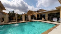The pool area is a well-appointed outdoor recreational space designed to provide a resort-like experience. The centerpiece is a generously sized swimming pool with clear turquoise water, complemented by an adjacent spa/hot tub for additional relaxation options. A standout feature of the pool is its decorative water elements — tall stone pillars with cascading waterfall spouts that add both visual interest and a soothing ambient sound to the environment.

The surrounding sundeck is finished in a light concrete surface, providing ample space for the numerous lounge chairs and seating arrangements positioned around the pool's perimeter. Shade umbrellas are stationed throughout the deck, offering relief from direct sun exposure.

A dedicated pool house anchors the space, constructed with stucco exterior walls, a pitched metal and shingle roof combination, and large glass windows and doors that provide direct views of the pool. Entry to the pool area is managed through a secure gated access point, enhancing safety and privacy. Decorative wood-framed doors and wall-mounted lighting add architectural character to the pool house entry.

The entire area is enclosed by black metal fencing, maintaining a sense of seclusion while remaining visually open to the surrounding landscape and mature trees overhead.