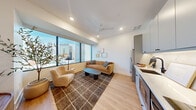 This thoughtfully designed urban residence occupies a single floor and showcases a clean, contemporary aesthetic throughout. Light oak hardwood flooring runs continuously across the space, unifying each area with warmth and cohesion.

A long, well-lit hallway with recessed lighting and crisp white walls — accented by a sliding barn door — welcomes you into the unit. It opens into a bright, open-plan living and kitchen area anchored by expansive floor-to-ceiling windows that frame sweeping city skyline views, flooding the space with abundant natural light. The living area features a ceiling fan and recessed lighting overhead, with ample room for comfortable seating arrangements.

The kitchen is efficiently designed with soft grey cabinetry extending to the ceiling, stainless steel appliances including a French door refrigerator and dishwasher, and light stone countertops — all contributing to a sleek, functional layout.

The bedroom is a calm, understated retreat with recessed lighting, a high clerestory window, and warm, neutral finishes that promote restful living. The bathroom continues the minimalist theme with large-format tile flooring, a glass-enclosed shower, a wood-toned vanity, and matte black fixtures that add a refined, modern contrast throughout.