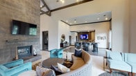 Located in Frisco, this single-story space makes a striking impression with its soaring vaulted ceilings accented by dark exposed wood beams, creating an open and airy atmosphere. The main living area is anchored by a sleek floor-to-ceiling stone fireplace on one wall, paired with a mounted flat-screen television, while bold teal accent chairs and a curved neutral sectional sofa arranged around a circular coffee table establish a sophisticated yet welcoming seating arrangement. Patterned area rugs add visual texture to the light-toned tile flooring throughout.

A seamless transition leads to a dedicated bar and lounge area at the rear of the space, complete with high-top bar seating, gray upholstered stools, and a decorative tile backsplash framing a second mounted screen. Recessed lighting illuminates the space alongside the natural light that filters through the open layout. Decorative wall accents, including a sunburst mirror and circular metallic wall art, contribute to the contemporary aesthetic. The overall design blends modern architectural elements with carefully curated furnishings, resulting in a refined, multi-functional environment ideal for both social gatherings and relaxed lounging.