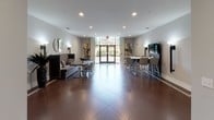 Located at 430 Allied Place in Gaithersburg, Maryland, this single-floor community lounge is a well-appointed gathering space designed for both casual socializing and collaborative use. The layout is open and spacious, featuring large-format tile flooring in a warm terracotta tone that anchors the room with a clean, contemporary aesthetic.

The main area is organized into distinct seating zones. A plush, high-backed booth-style sofa with decorative pillows provides a comfortable lounge nook, accompanied by a wooden dining table and sleek modern chairs. Opposite, a tall bar-height white table with contemporary stools serves as a secondary gathering spot, centered beneath a wall-mounted flat-screen television flanked by stylish sconce lighting. A smaller round table with dark chairs offers additional intimate seating.

Natural light filters through large glass double doors at the entrance, which frame views of the surrounding greenery. Contemporary abstract artwork adorns the walls, adding visual interest alongside the warm, ambient wall sconces. The color palette throughout is neutral and modern, with soft gray walls providing a clean backdrop.

A separate corridor leads to restroom facilities on either side, with clear signage and a mirrored view back toward the main entrance. The overall ambiance is polished and inviting, making this lounge an ideal multipurpose space for community gatherings and everyday use.