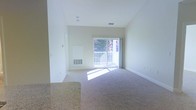 This single-floor residential space offers a clean, functional layout designed for comfortable everyday living. The main living area is finished with soft, plush carpeting and bathed in natural light that streams through a full sliding glass door, which opens directly onto a private balcony with a white railing. The outdoor views feature lush, mature trees, bringing a sense of nature and tranquility into the interior. The walls are painted in a crisp, neutral white tone, lending the space an airy and open feel. A floor-mounted HVAC vent ensures year-round climate comfort, while a ceiling light fixture provides overhead illumination. Vaulted or elevated ceilings enhance the sense of vertical space, making the living area feel particularly open and unconfined. A partial view of a granite countertop surface is visible, hinting at a kitchen or dining area that transitions seamlessly into the living space. The overall ambiance is calm and light-filled, with a neutral color palette that provides a versatile backdrop ready for personalization. The unit presents a welcoming, move-in-ready environment with thoughtful details throughout.
