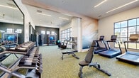 This well-appointed fitness center at 1500 Westshore Dr in Cumming, Georgia, offers a comprehensive workout environment designed with both functionality and aesthetics in mind. The single-floor facility is organized into distinct training zones, each thoughtfully equipped to support a variety of fitness routines. The cardio area features treadmills, elliptical machines, stationary bikes, and a WaterRower, all positioned alongside large grid-pane windows that flood the space with natural light and offer views of the surrounding outdoors. The strength training section includes a full dumbbell rack, adjustable weight benches, and cable/functional training machines, providing options for users of all fitness levels. A dedicated functional training area is outfitted with resistance bands, kettlebells, medicine balls, stability balls, and a multi-use rig, complemented by floor-to-ceiling mirrors that enhance the sense of space. The interior design is elevated by a striking abstract wall mural rendered in soft ocean-inspired tones, contrasting beautifully with the dark accent walls. Recessed linear lighting and ceiling fans maintain a bright, comfortable atmosphere throughout. Patterned carpet flooring ties the space together, adding a modern, polished finish to this fully equipped community fitness facility.