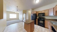 This two-floor residence offers a practical and comfortable layout designed for everyday living. The heart of the home is an open-concept kitchen and living area, where light tile flooring flows seamlessly through the space. The kitchen is equipped with black appliances, including a refrigerator, range, and built-in microwave, all complemented by warm maple-toned cabinetry that provides ample storage. A central kitchen island with a dark countertop offers additional workspace and a natural gathering point. A pendant chandelier with warm-toned lighting hangs above the transition between the kitchen and the carpeted living area, adding a welcoming touch. The textured ceiling adds subtle dimension to the space. Natural light filters in through a window in the living area, brightening the open layout. A doorway visible from the main living space leads to additional rooms, suggesting a well-connected floor plan. The combination of tile and carpet flooring defines the distinct functional zones while maintaining a cohesive aesthetic throughout. The two-floor layout provides additional space and versatility for a variety of living arrangements.