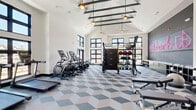 Located at 5617 Oakley Industrial Blvd in South Fulton, GA, this fitness center is a thoughtfully designed, single-floor facility that delivers a premium workout experience. The space is defined by its striking vaulted ceiling with exposed dark steel beams, which lend an industrial-chic aesthetic while creating an expansive, airy atmosphere. Pendant globe lights and ceiling fans complement the architectural detail above.

Large black-framed windows line the walls, flooding the interior with natural light and offering views of the surrounding outdoors. A set of double glass doors provides direct exterior access, further connecting the space to its environment. The geometric diamond-patterned carpet tile flooring in shades of blue, gray, and cream adds a contemporary visual energy throughout.

The fitness floor is comprehensively equipped for both cardio and strength training. Cardio offerings include treadmills, ellipticals, stationary cycling bikes, and a rowing machine. The strength training area features a power rack, adjustable benches, a full dumbbell rack, kettlebells, medicine balls, resistance cables, and a cable machine. A dedicated accent wall with neon signage adds a stylish, modern touch, while a wall-mounted display screen provides informational content. Towel storage and a stability ball round out the thoughtful amenities of this well-appointed facility.