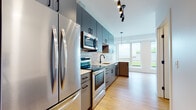 This single-floor unit showcases a contemporary design with consistent light wood-tone flooring flowing throughout the space. The open-concept kitchen is a standout feature, equipped with full stainless steel appliances including a French door refrigerator, electric range, built-in microwave, and dishwasher. Sleek gray cabinetry extends from floor to ceiling, complemented by matte black hardware and clean white countertops. A striking geometric triangle-patterned tile backsplash adds visual texture behind the sink and cooking area. Track lighting and a pendant fixture provide layered illumination. The kitchen opens directly to the living area, which benefits from large windows that frame outdoor views and draw in generous natural light. A dedicated bedroom area features an oversized window with sweeping exterior views and warm wood flooring. The bathroom includes a full soaking tub and shower combo with matte black fixtures, maintaining the modern aesthetic seen throughout. A spacious walk-in closet with wire shelving systems offers practical storage. The overall design is clean, modern, and cohesive, with neutral tones and contemporary finishes creating a polished, move-in-ready environment.