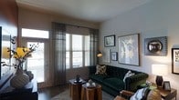 This single-floor space centers around a thoughtfully designed living room that balances comfort and style. The room receives generous natural light through a large window fitted with horizontal blinds and sheer, draped curtains in a neutral gray-green tone, which soften the incoming light while maintaining a sense of openness. A glass-paneled door provides direct access to an outdoor area, further enhancing the bright and airy atmosphere.

The focal point of the room is a deep emerald green tufted sofa, offering a rich, bold contrast against the neutral warm-beige walls. Patterned throw pillows in earthy tones complement the sofa's color palette. Two organic, raw-edged wooden side tables serve as functional coffee tables, grounding the space with a natural, rustic element that balances the more polished furnishings.

The walls are adorned with a curated selection of framed artwork, including a large abstract gold-and-white piece and smaller botanical and illustrative prints, adding visual interest and personality to the space. A floor lamp with a warm-toned shade provides ambient lighting. The flooring consists of dark hardwood, partially covered by a light-toned area rug, tying the room's design elements together cohesively.